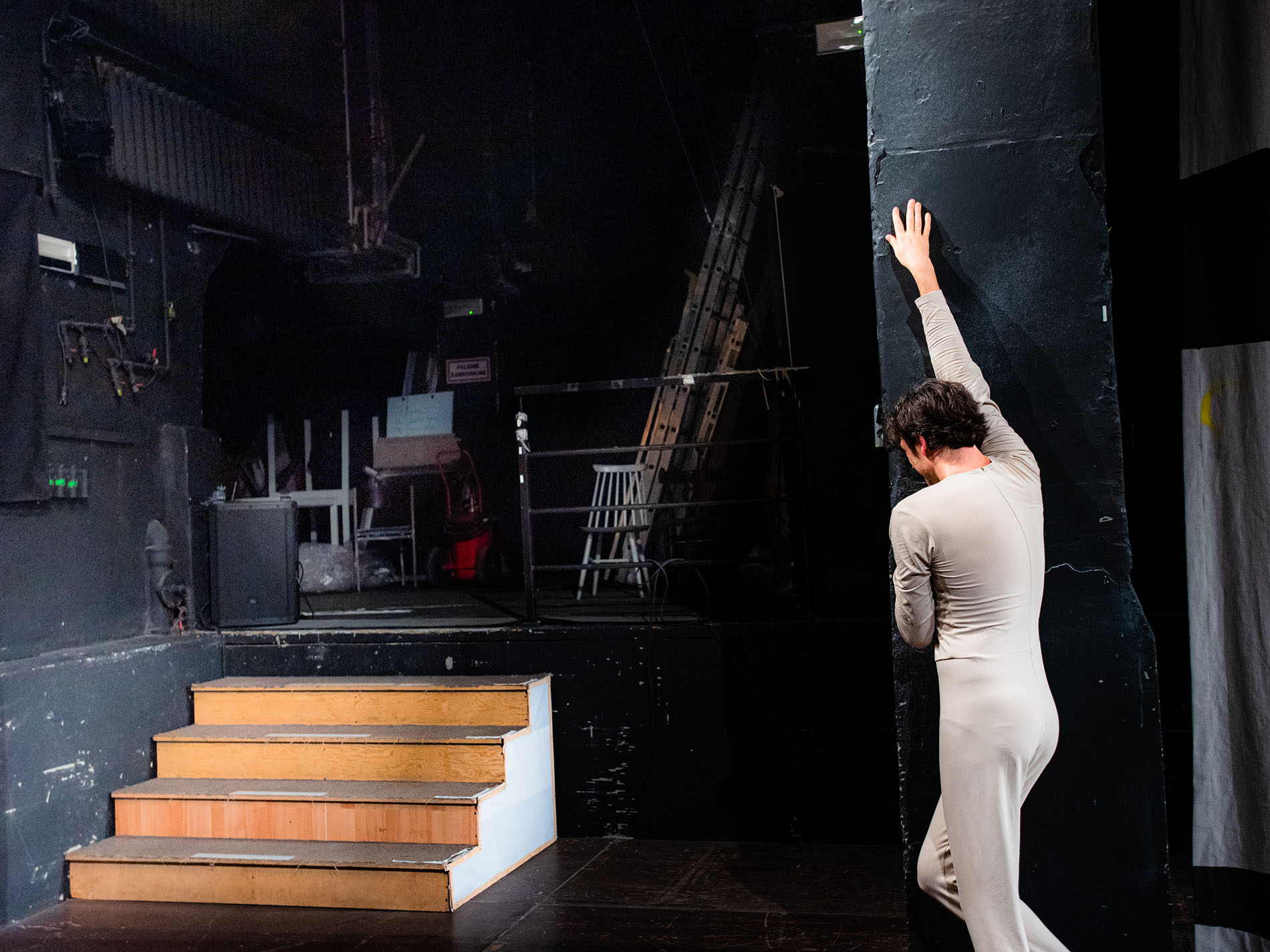 Po prawej, przy czarnej ścianie, ciemnowłosy mężczyzna w trykocie. Stoi tyłem. Po lewej drewniane schodki prowadzące na teatralne zaplecze.